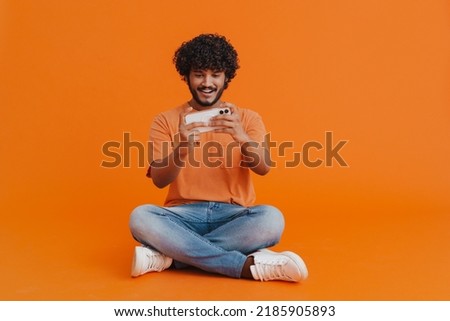 Young indian handsome curly man playing mobile game sitting in lotus pose over isolated orange background