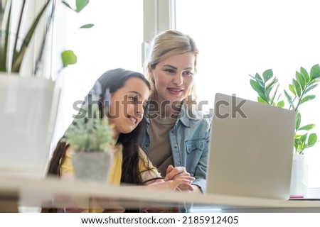 Beautiful mother and child looks at laptop screen spend time in kitchen using app education program, choose cartoons, teach kid pc usage, controls what daughter watching on internet cyberspace