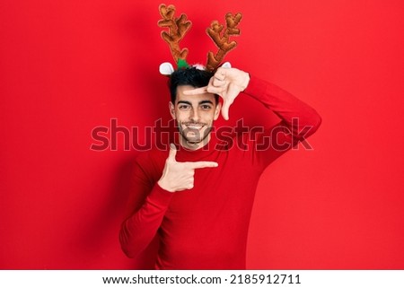 Young hispanic man wearing cute christmas reindeer horns smiling making frame with hands and fingers with happy face. creativity and photography concept. 