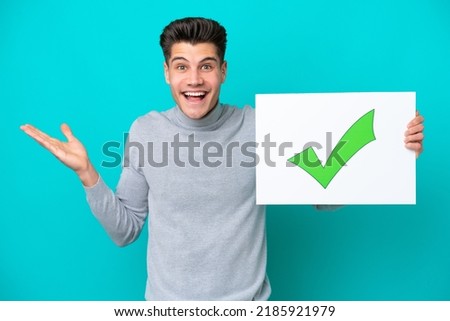 Young handsome caucasian man isolated on blue bakcground holding a placard with text Green check mark icon with surprised expression
