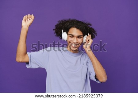 Young happy smiling handsome latin curly man in headphones with closed eyes dancing and enjoying music over isolated violet background