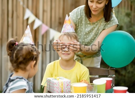 Mom from behind closes eyes her son with hands. Cute funny nine year old boy celebrating his birthday with family and friends with homemade baked cake in a backyard. Birthday party