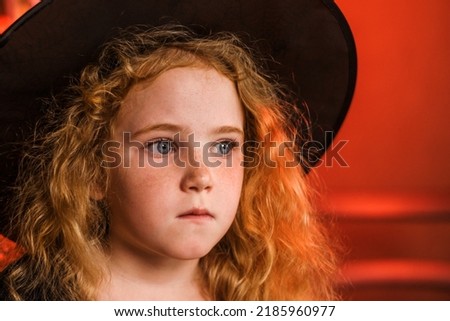 Portrait of cute red-haired girl in a black witch costume and hat on an orange background.Halloween concept.Selective focus,close-up.