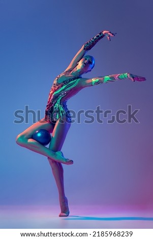 Portrait of young beautiful girl, rhythmic gymnast athlete posing with blue ball isolated on blue purple background in neon light. Concept of action, motion, sport life, motivation, competition.
