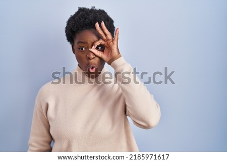 African american woman standing over blue background doing ok gesture shocked with surprised face, eye looking through fingers. unbelieving expression. 