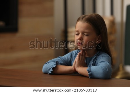 Cute little girl with hands clasped together praying at table indoors Royalty-Free Stock Photo #2185983319