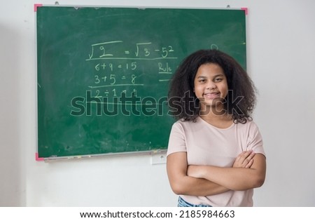Girl is standing  in a classroom with a green chalkboard covered with formulas and figures. Kid write on chalk board.