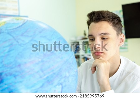 Portrait of a thoughtful young man in a white shirt sitting at a table in front of a globe. The concept of geography lesson, travel. High quality photo