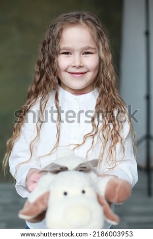 Blond Caucasian female child with dark narrow eyes indoors holding soft toy and looking cheerfully smiling. Wearing white t-shirt. Studio portrait. Happy childhood concept. Candid authentic image Royalty-Free Stock Photo #2186003953