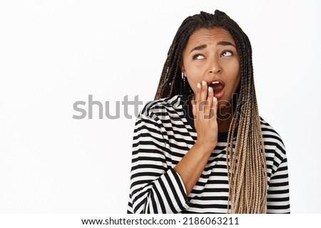 Close up portrait of black girl yawning, looking aside with unamused bored face expression, standing in striped blouse over white background