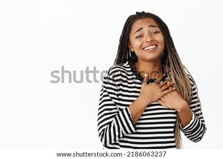 Portrait of beautiful black girl looking with yearning and tenderness, showing heartbeat gesture, holding hands on heart and smiling, standing over white background Royalty-Free Stock Photo #2186063237