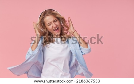 Young attractive girl has fun listening to loud music in headphones and dancing in casual clothes on a pink background