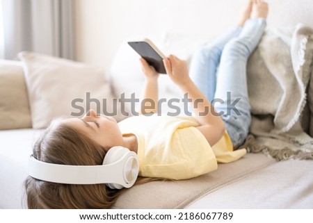 girl lying at home on cozy sofa dressed casual jeans listening to music and watching cartoons using wireless headphones connected with smartphone. 