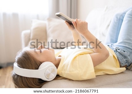 girl lying at home on cozy sofa dressed casual jeans listening to music and watching cartoons using wireless headphones connected with smartphone. 