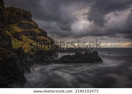 Rock and water lines in a black sand beach with moody cliffs on the background in Seixal at stormy day. October 2021. Madeira, Portugal. Long exposure picture.