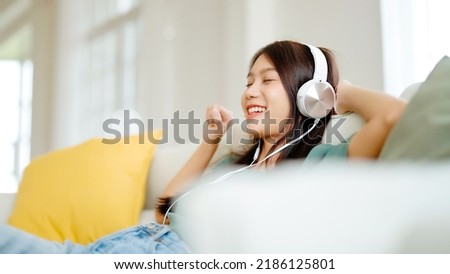 Young asian woman listening to music on couch in living room at home. Happy asia female using mobile smartphone, wearing headset and sitting on sofa Royalty-Free Stock Photo #2186125801
