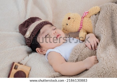 Asian adorable newborn baby wear brown knit hat deeply sleeping with beige blanket next to teddy bear and toy camera with safe and comfortable.  Little toddler cute resting after eat full.