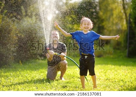 Funny little boy with his father playing with garden hose in sunny backyard. Preschooler child having fun with spray of water. Summer outdoors activity for family with kids. Dad involved in parenting Royalty-Free Stock Photo #2186137081