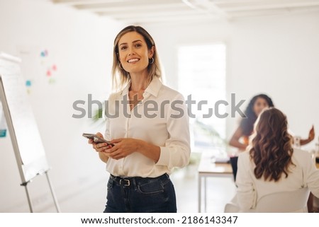 Happy young businesswoman smiling at the camera while holding a smartphone in a meeting room. Cheerful young businesswoman working in an all-female office. Royalty-Free Stock Photo #2186143347
