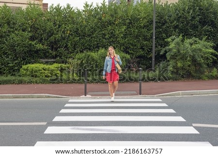 A girl with fair hair in a red dress and a denim jacket with a bag on her shoulder crosses the road at a pedestrian crossing. The concept of traffic rules Royalty-Free Stock Photo #2186163757