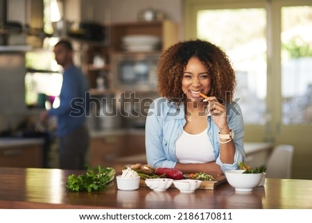 When it comes to our food we keep it clean. Portrait of a young woman eating a carrot while preparing a healthy meal with her husband in the background. Royalty-Free Stock Photo #2186170811