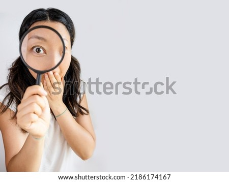Funny image of young surprised asian woman looking at camera through magnifying glass, isolated on white background