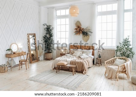 Cozy rustic bedroom with boho ethnic decor. Bright spacious apartment with large windows. Nobody. Wooden furniture. Boudoir table. Large mirror. Handmade textile. Plants in the interior. Royalty-Free Stock Photo #2186209081