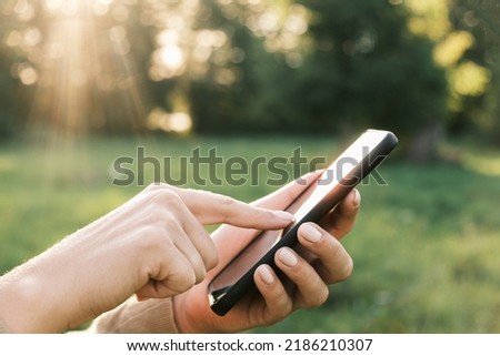 Closeup of female hands using a smart phone, nature background. Woman using mobile phone in sunset outdoors. Unrecognizable person touching smartphone screen. Unknown girl holding cellphone outside.