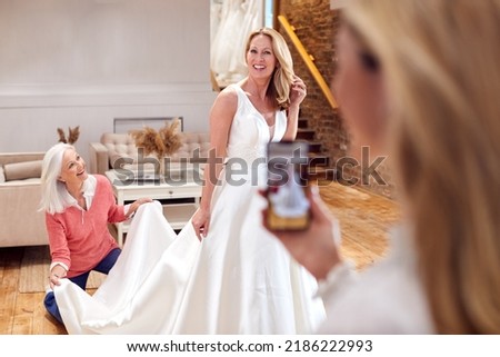 Grandmother With Adult Daughter And Granddaughter Trying On Wedding Dress Taking Picture On Phone