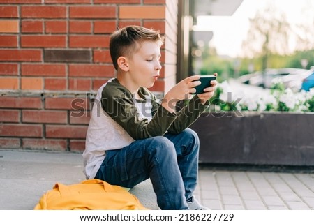 Back to school. Cute child with backpack, holding mobile phone, playing with cellphone. School boy pupil with bag. Elementary school student after classes. Kid sitting on stairs outdoors in the Royalty-Free Stock Photo #2186227729