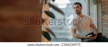 A young man sits on a windowsill and looks at the city on a rainy day. A handsome, pumped up brunette guy sits thoughtfully by a large window at his home on a cloudy day. Royalty-Free Stock Photo #2186236975