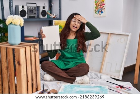 Hispanic young woman holding notebook at art studio stressed and frustrated with hand on head, surprised and angry face 