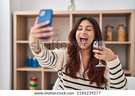 Hispanic young business woman taking a selfie picture drinking a glass of wine smiling and laughing hard out loud because funny crazy joke. 