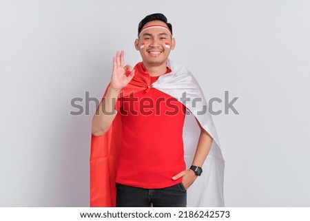 Happy young Asian man holding the Indonesian flag while showing okay gestures isolated on white background. Indonesian independence day concept