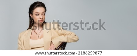 brunette young woman in stylish outfit looking away and leaning on chair isolated on grey, banner Royalty-Free Stock Photo #2186259877