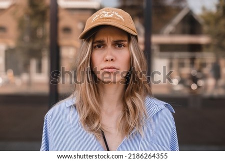 Drama blonde curly woman look sad at camera walking on the street. Girl wear beige cap and stripped shirt. Portrait of offended young girl with pouty lips wearing casual clothing standing. Royalty-Free Stock Photo #2186264355