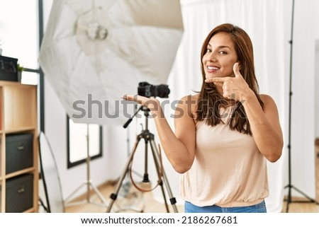 Young latin woman standing at photography studio amazed and smiling to the camera while presenting with hand and pointing with finger. 