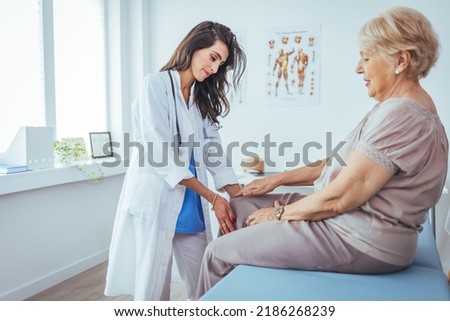 Pain in the knee. Good looking nice aged woman holding her knee and looking at her doctor while explaining where she feels pain. Support for strained muscles. Doctor fixing womans knee with hands