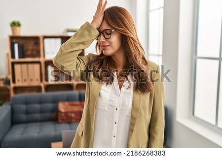 Hispanic woman working at consultation office surprised with hand on head for mistake, remember error. forgot, bad memory concept.  Royalty-Free Stock Photo #2186268383