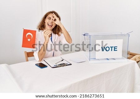 Beautiful caucasian woman at political campaign election holding tunisia flag smiling happy doing ok sign with hand on eye looking through fingers 