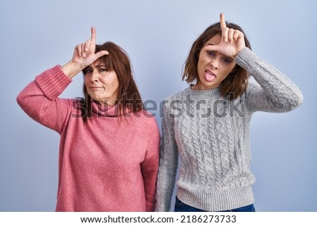 Mother and daughter standing over blue background making fun of people with fingers on forehead doing loser gesture mocking and insulting. 