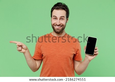 Young smiling happy man 20s wear orange t-shirt hold in hand use mobile cell phone with blank screen workspace area point index finger aside isolated on plain pastel light green background studio.
