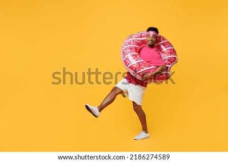 Full body side view young man he wear pink t-shirt bandana near hotel pool hold inflatable rubber ring raise up leg leaning back isolated on plain yellow background. Summer vacation sea rest concept