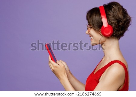 Side view young smiling woman 20s she wear red tank shirt eyeglasses headphones listen to music hold in hand use mobile cell phone isolated on plain purple background studio. People lifestyle concept Royalty-Free Stock Photo #2186274665