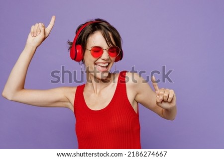 Young happy cheerful woman 20s she wear red tank shirt eyeglasses headphones listen to music dance point index fingers up isolated on plain purple background studio portrait People lifestyle concept