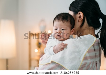 closeup innocent asian infant baby leaning on her mom’s shoulder with cloth is looking away into the space while her mom is burping patting on her back at home. Royalty-Free Stock Photo #2186286965