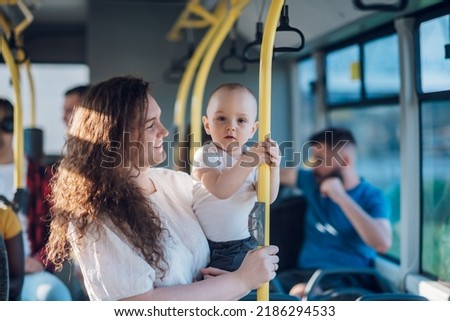 Mother carries her child while standing and holding on to the bus.Mom holding her infant baby boy in her arms while riding in a public transportation. Cute toddler boy traveling with his mother. Royalty-Free Stock Photo #2186294533