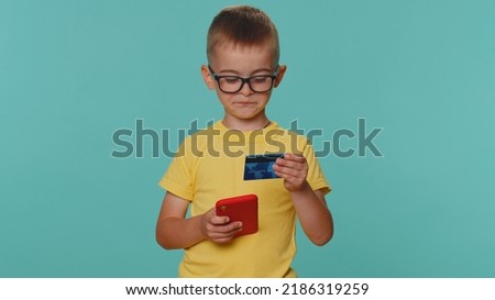 Little toddler children boy in yellow t-shirt using credit bank card and smartphone while transferring money, purchases online shopping. Young preschool kid isolated alone on blue studio background