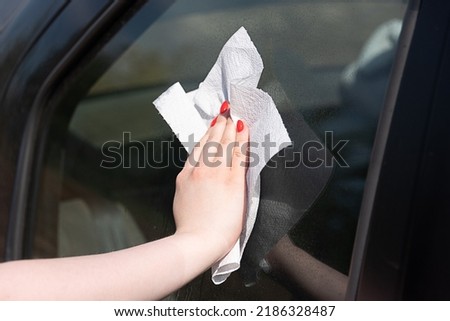 Woman wipes car glass with paper napkin, glass in spray drops, reflection on glass. Car service outdoors on a summer day.