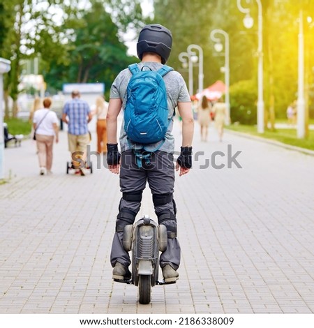 Man on motorized unicycle riding trough the city park in summer day. Man cycling on monowheel in protective gear. Man with backpack wear helmet, racing gloves, knee protection rides on single wheel Royalty-Free Stock Photo #2186338009